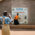 Eine Schülerin aus Uganda steht vor einer Wand mit einer Sehtafel. Sie versucht die Buchstaben zu sehen, auf die eine Frau in einem orangen Shirt deutet.