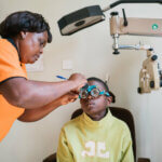 Justine Basicila is is sitting on a chair and getting his eyes examined by a woman wearhing an orange shirt.