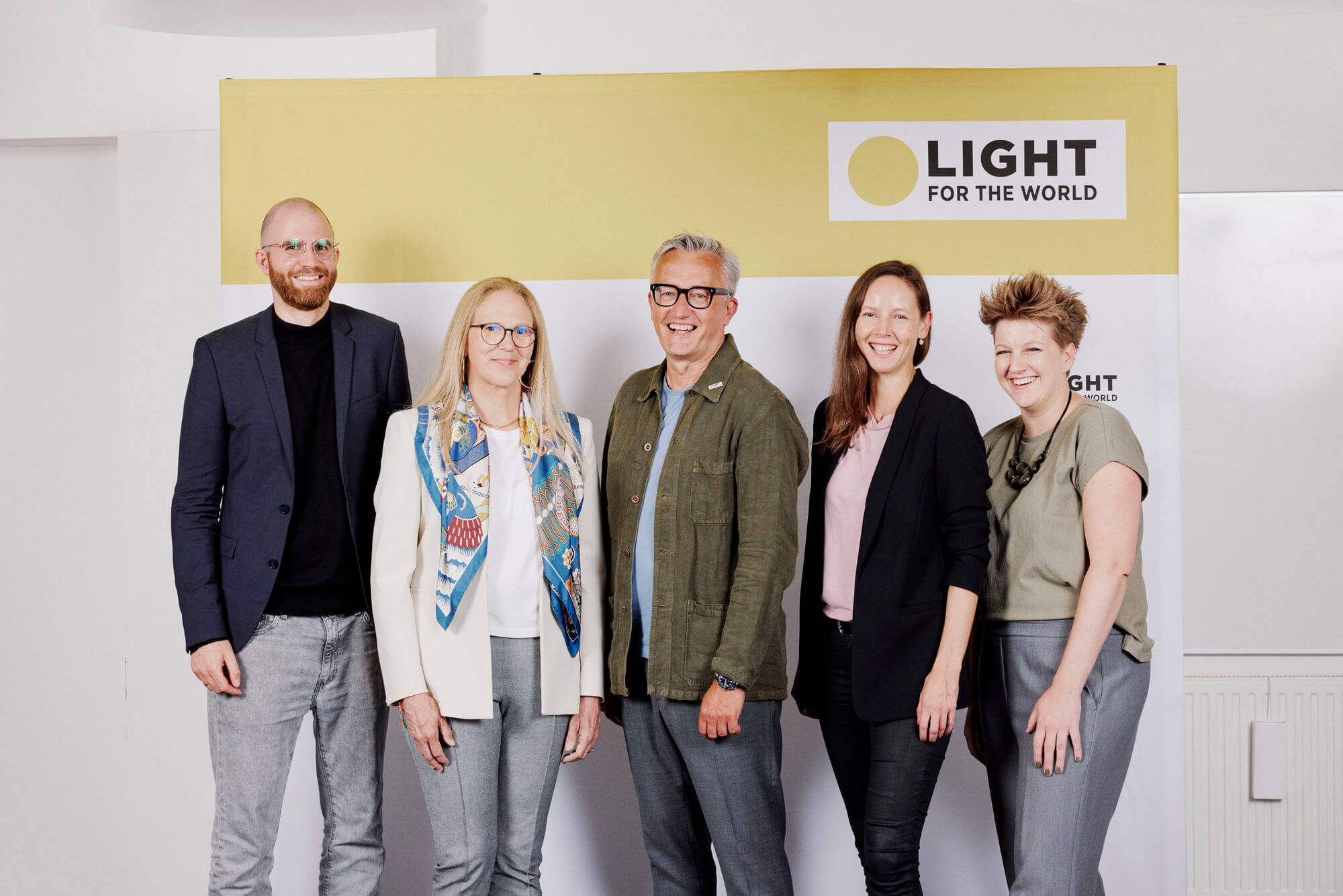 Das Internationale Managementteam von Licht für die Welt steht nebeneinander vor einer Logowand und lächelt in die Kamera.