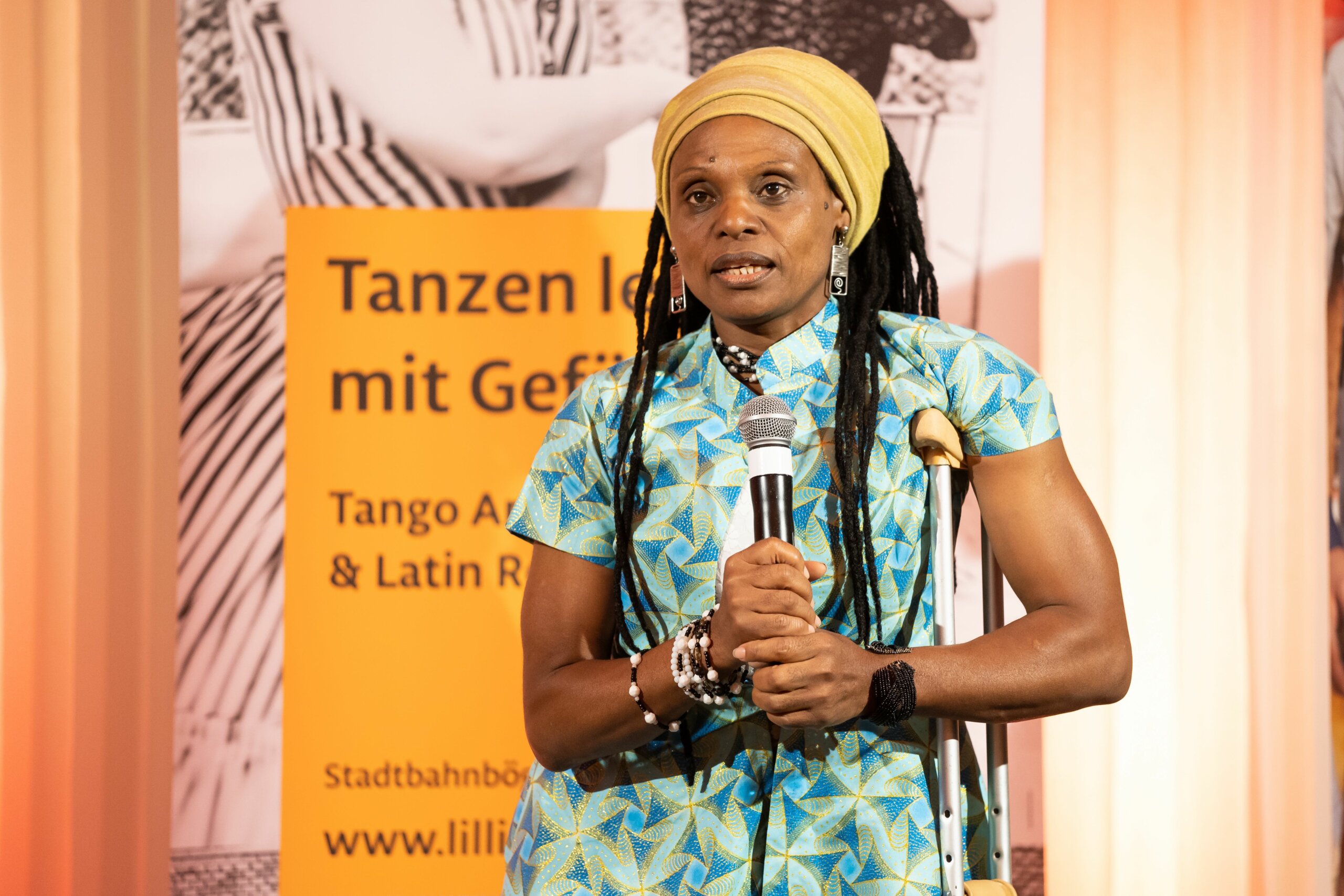 An African woman with long black hair and yellow scarf on the head holds a microphone. She is in light green and white dress