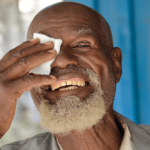 Ein älterer Herr mit weißem Bart drückt ein weißes Tuch auf sein frisch operiertes Auge. Er lächelt breit in die Kamera.