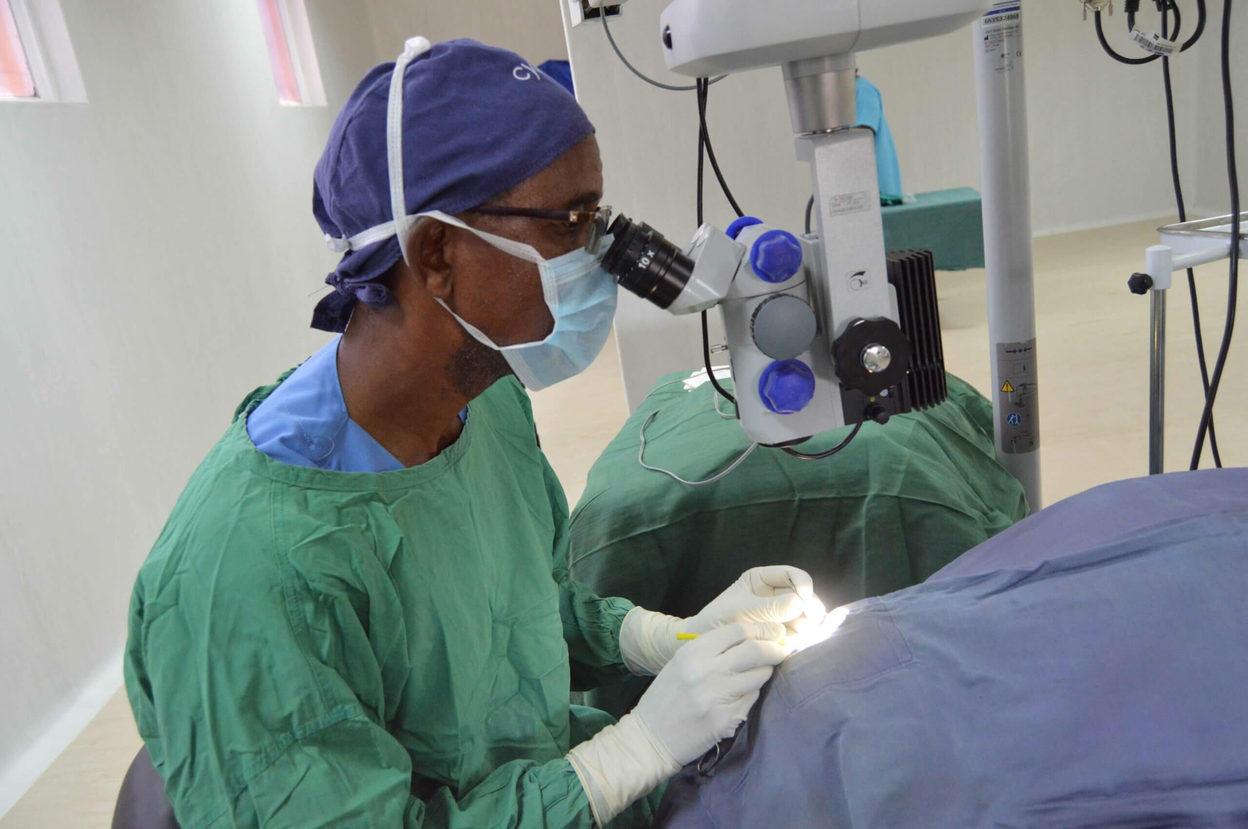 Dr. Abel Polaze aus Mosambik blickt durch ein Gerät, während er eine Person vor sich am Tisch operiert.