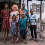 Eine Familie steht vor ihrem Haus und lächelt in die Kamera. Die Mutter, umringt von ihrer Familie, lebt mit Albinismus. Sie hat blonde Haare und trägt ein beige-weißes Kleid.