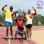 Drei Spitzensportler mit Behinderungen stehen am Sportplatz, die Hände jubelnd in die Luft gestreckt und lächeln in die Kamera. Alle tragen ihre Sportbekleidung.