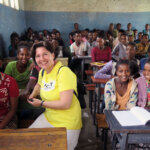 Geschäftsführerin von Light for the World Deutschland Astrid Pietig sitzt in einem Klassenzimmer in Äthiopien. Sie trägt ein gelbes Shirt von Light for the World und um sie herum sitzen die Schulkinder in ihren Schulbänken.