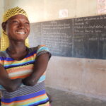 Eine junge Frau mit einem gestreiften T-Shirt und Armen, die an den Ellenbogen enden lächelt in die Kamera. Sie steht in einem Klassenzimmer.