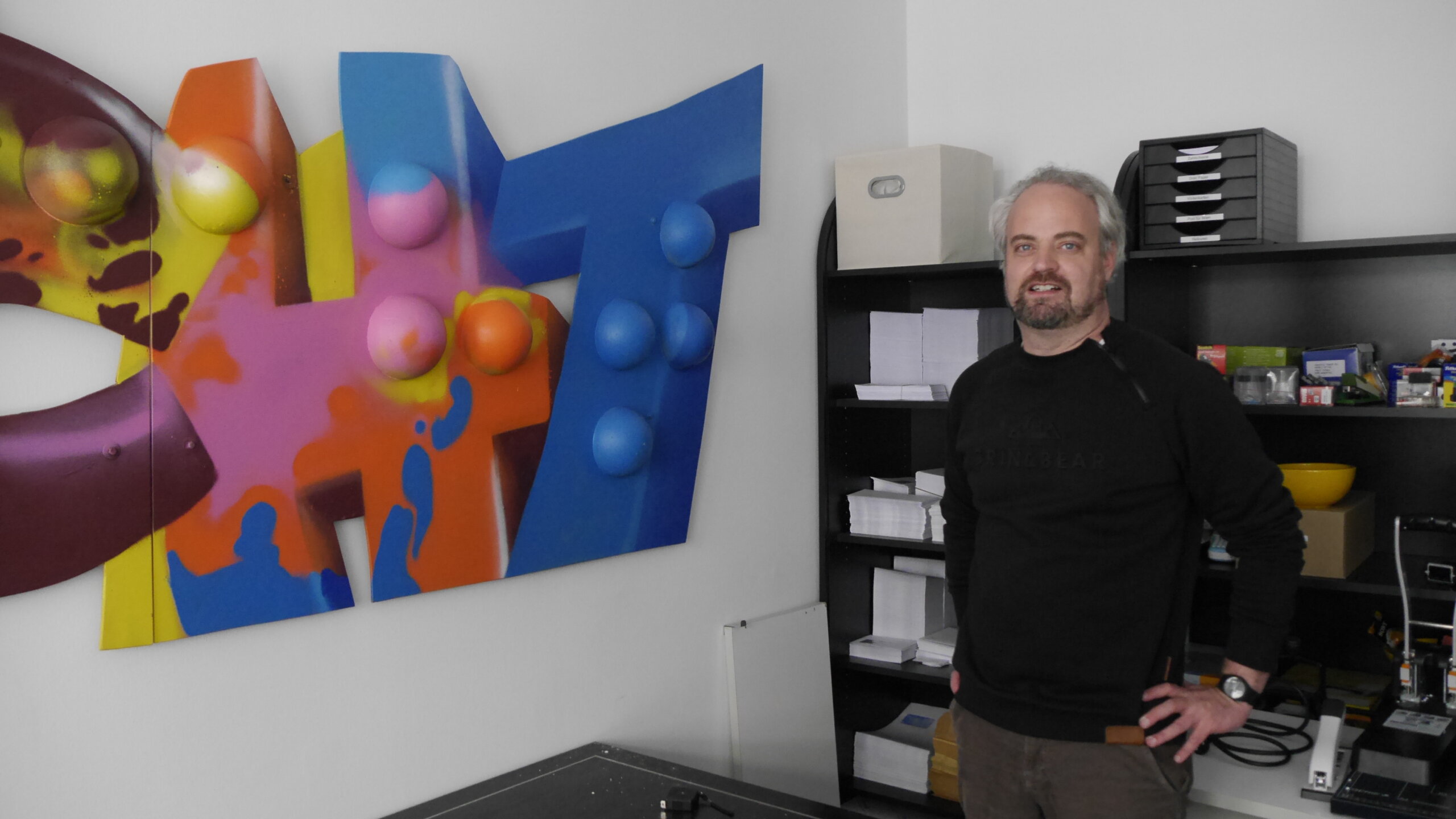 Ehrenamtlicher Martin Kitasaka steht nach getanter Arbeit mit Händen auf die Hüften gestemmt im Büro neben dem eben angebrachten Graffiti-Braille-Kunstwerk "Licht"