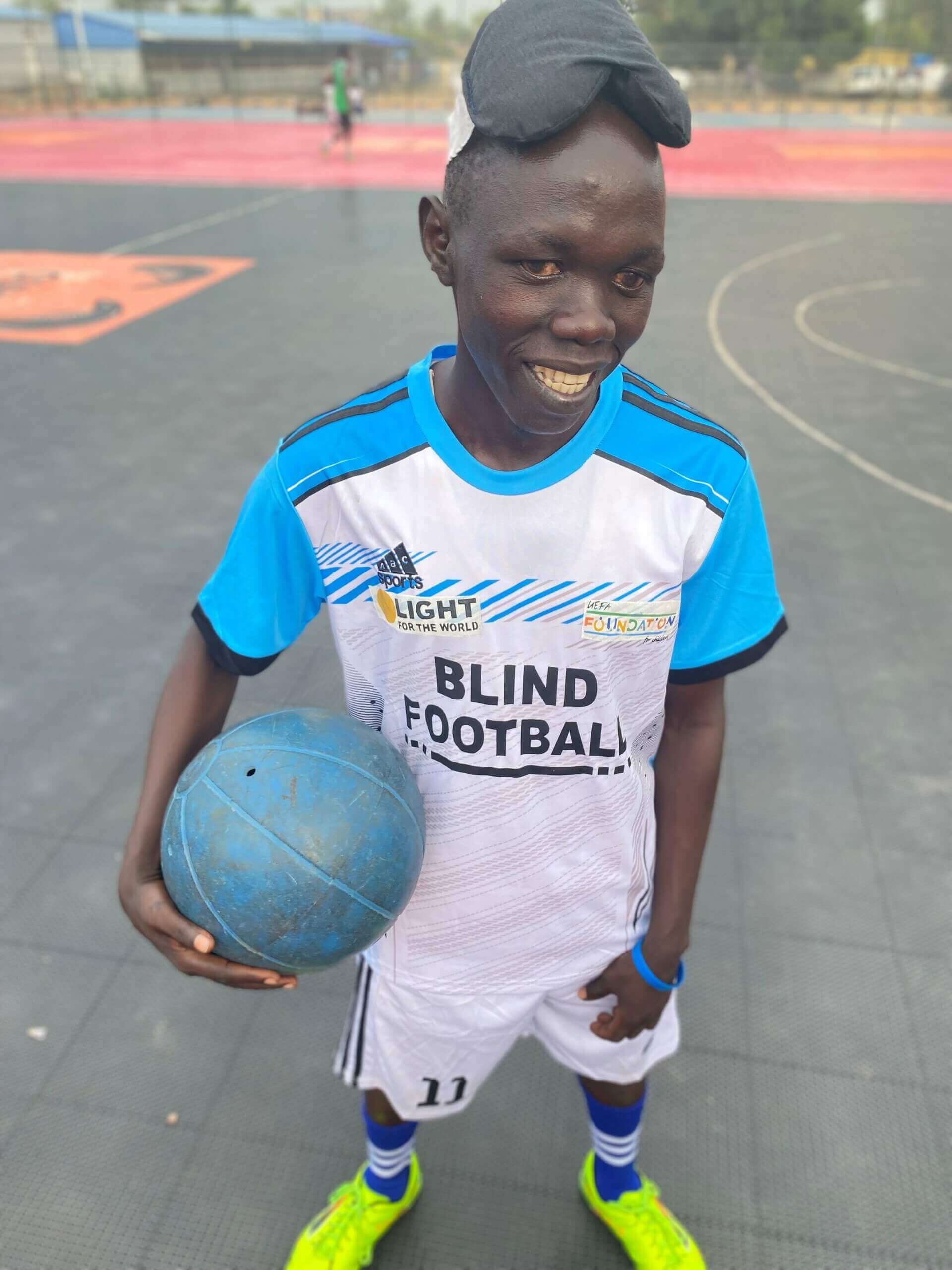 Jimmy ist Teil des Blindenfußballteams im Südsudan. Er trägt ein weißblaues Trikot, hält den Ball in seinem linken Arm und lächelt in die Kamera. Hinter ihm ist das Fußballfeld zu sehen.