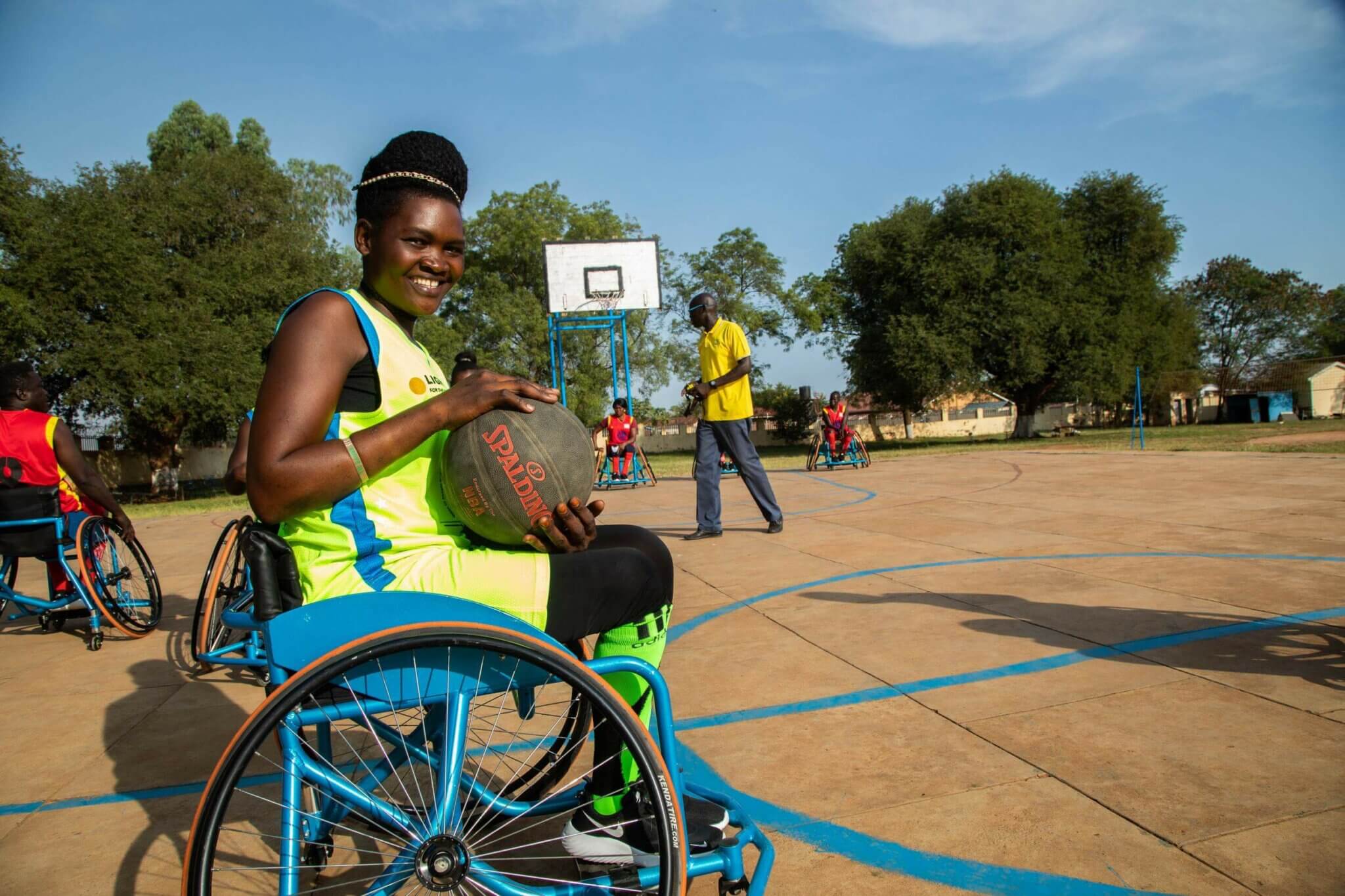 Lucy, eine Inklusionsberaterin von Licht für die Welt in Mosambik, sitzt im Rollstuhl und nimmt an einem Basketballtrunier teil. Sie trägt ein gelbes Trikot und hält den Ball lächelnd in ihren Händen.