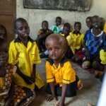 Die vierjährige Nyamush sitzt am Boden im Südsudan umringt von ihren Schulkolleg*innen. Alle Kinder tragen eine gelbe Bluse mit schwarzem Kragen und schwarze Hosen. Ihr Blick ist wach und interessiert nach oben gerichtet.