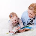Eine blonde Mutter liegt mit ihrem kleinen Kind auf einem weißen Fellteppich und zeigt ihm ein Bilderbuch. Das blonde Kind blickt begeistert in das Buch hinein. Beide lachen.