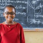 Ein Mädchen, das eine Brille und ein rotes Oberteil trägt, steht lächelnd vor einer Tafel in einem Klassenzimmer.
