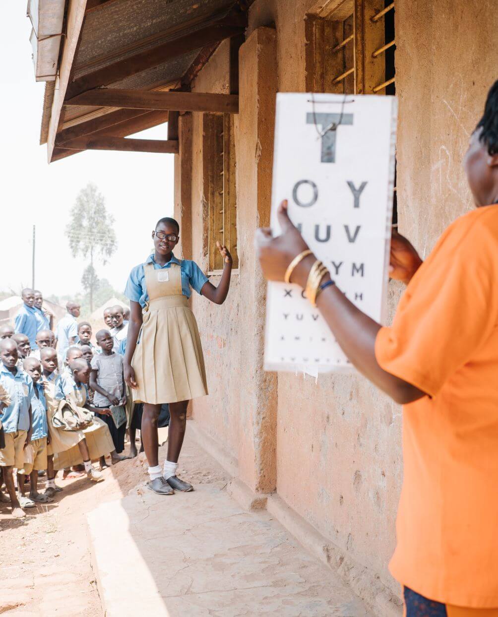 Sehtest an einer Schule in Uganda. Eine Frau in einem orangen T-Shirt hält eine Tafel hoch, auf der Buchstaben in unterschiedlichen Größen zu sehen sind. Eine Schülerin, die etwas entfernt davon steht, liest die Buchstaben vor.