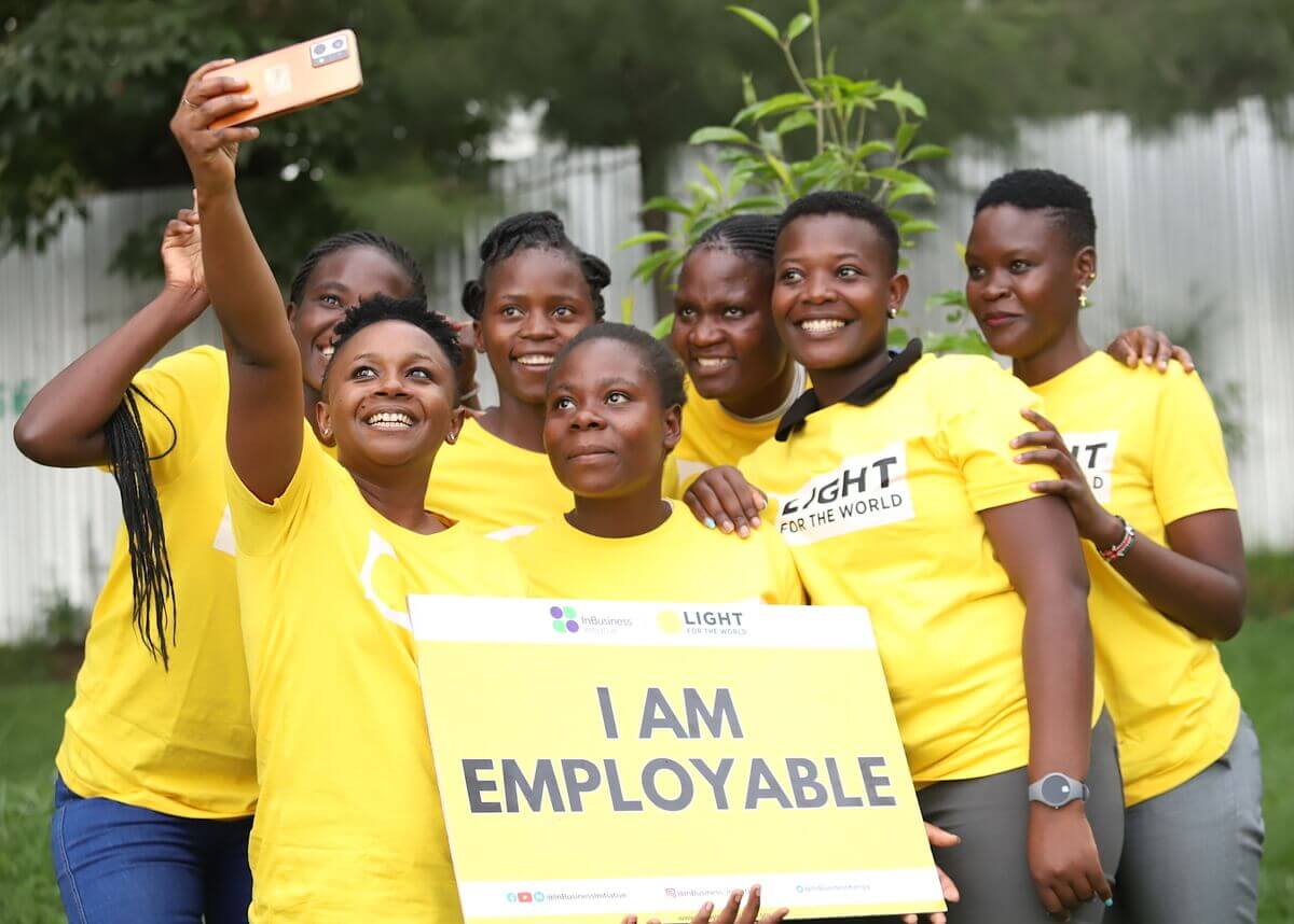 Sieben Frauen im gelben Light for the World T-Shirt posieren für ein Foto mit dem Schild "I am employable"