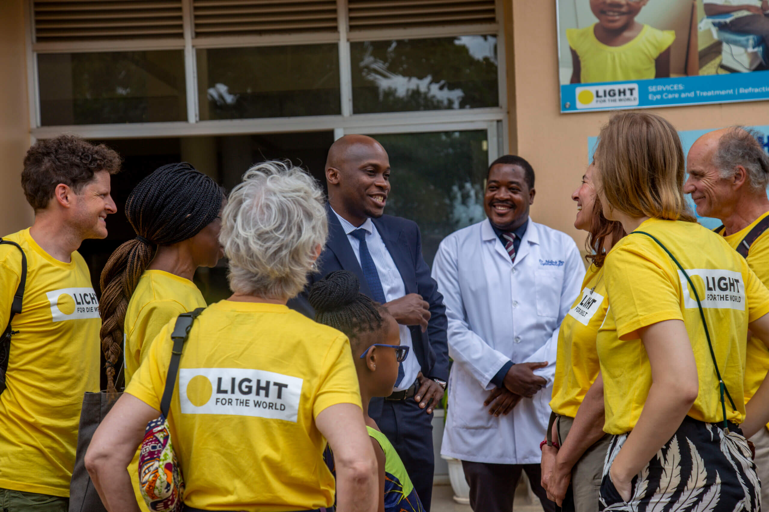 Eine Gruppe von Augenärzt*innen aus Deutschland, Österreich und der Schweiz auf einer Projektreise steht mit zwei Herren vor dem Mulago Hospital in Uganda. Sie erhalten gerade Informationen über das Krankenhaus.