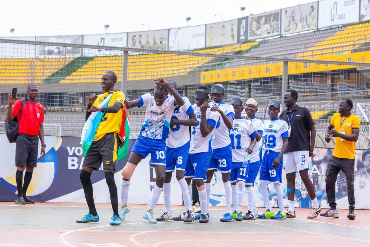 Auf dem Bild ist die südsudanesische Blindenfußballmannschaft zu sehen. Sie tanzen, weil sie die Afrika-Meisterschaft gewonnen haben. Sie tragen blau -weiße Trikots. Der Tormann ganz vorne trägt ein gelb-schwarzes Trikot.
