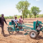 Das Bild ist auf einem leeren Feld aufgenommen. Eine Frau mit einer Behinderung sitzt auf einem Pluggerät, während zwei schwarze Männer neben ihr hergehen. Das Wetter ist sonnig, der Himmel blau.