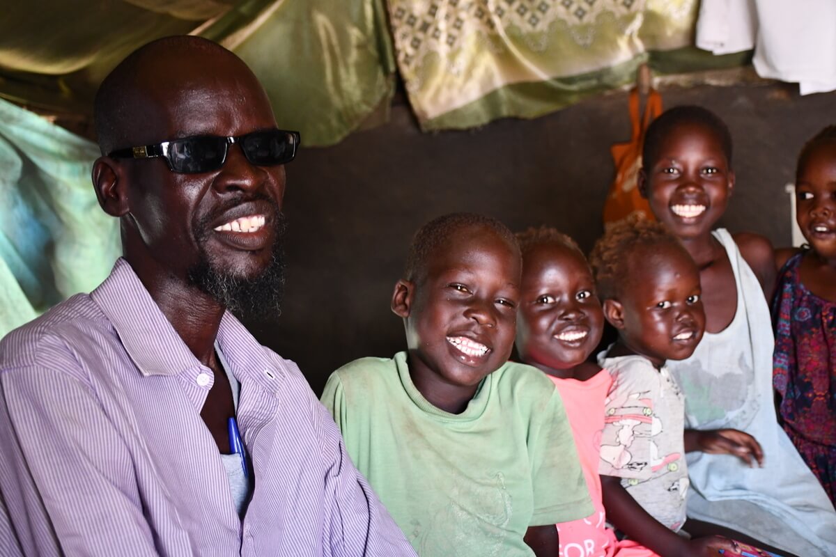 Auf dem Foto sind 6 Perosnen mit schwarzer Hautfarbe zu sehen, 1 erwachsener Mann und 5 Kinder. Sie alle lächeln in die Kamera. Das Bild wurde in einem Camp für geflüchtete Menschen aufgenommen.