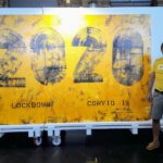 Ein Unterstützer von Licht für die Welt steht in einem gelben T-Shirt vor einem Plakat der Kunst für Covid-Hilfe in Afrika