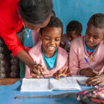 Ein Kind mit Behinderung sitzt lachend neben seinen Klassenkameradinnen. Sie tragen alle einen rosa Pulli. Die Lehrerin beugt sich über das Kind und hält seine Hand, während es in dem Heft vor ihm zu schreiben versucht.
