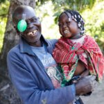 Franciscu aus Mosambik hatte gerade eine Operation am Grauen Star. Er hält seine lachende Enkelin in dne Armen und blickt lachend in die Kamera.