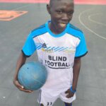 Jimmy ist Teil des Blindenfußballteams im Südsudan. Er trägt ein weißblaues Trikot, hält den Ball in seinem linken Arm und lächelt in die Kamera. Hinter ihm ist das Fußballfeld zu sehen.
