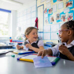 Zwei Mädchen sitzen an einem Tisch in der Schule und lächeln sich an. Beide tragen Schuluniformen, haben einen Stift in der Hand und ein Schreibbrett vor sich.