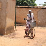 A woman with a disability that impairs her ability to walk, rides a bicycle while balancing her cane over the bike handles.