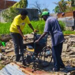 Musa aus Mosambik lehnt an seinem Rollstuhl, den eine Mitarbeiterin von Licht für die Welt vorbereitet hat. Sie stehen auf Musas Grundstück. Im Hintergrund sind Trümmer von Musas Haus zu sehen, das zur Gänze vom Wirbelsturm Idai 2019 zerstört wurde.