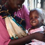 Auf dem Bild sind Nyakhan und ihre Mutter Nawal aus dem Südsudan abgebildet. Nawal hält ihre Tochter, die mit der Hirnkrankheit Hydrozephalus lebt, im Arm und lacht sie an. Nyakhan hat ein breites Grinsen im Gesicht.