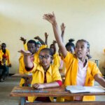 Der Bildausschnitt zeigt Nigest Uche aus ihrer Klasse in Äthiopien und ihre Schulkolleg*innen. Sie alle sitzen jeweils zu zweit an einem Holztisch und zeigen gerade auf.