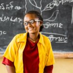 Nigest aus Äthiopien steht in ihrem Klassenzimmer vor der beschriebenen Tagel und lächelt. Sie trägt einen roten Pullover mit einem gelben Hemd darüber, welches Teil der Schuluniform ist.