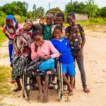 Teresa laughs outside, with 8 of her friends standing behind her. She sits in a wheelchair and holds her schoolbags and pencils.