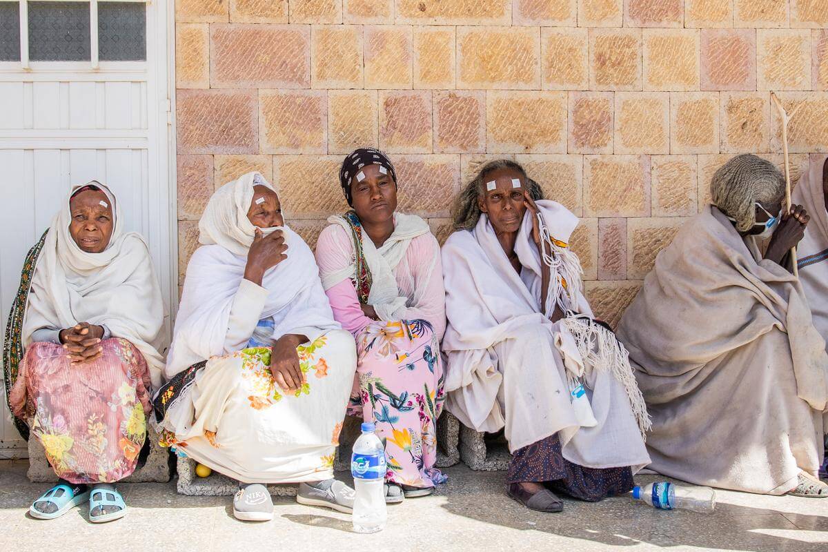 fünf Frauen sin hellen Gewändern sitzen in einer Reihe auf dem Boden und warten auf eine augenmedizinische Behandlung