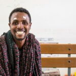Haile Tesfaye, ein 32-jähriger Mann, sitzt auf einer Bank und lächelt strahlend in die Kamera. Über seinen Augen hat er Markierungen.