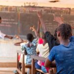 Auf dem Bild ist eine Klasse zum Schulanfang von hinten zu sehen. Die Schülerinnen und Schüler zeigen auf, während der Lehrer etwas an der Tafel erklärt. Das Bild entstand an einer Schule in Burkina Faso.