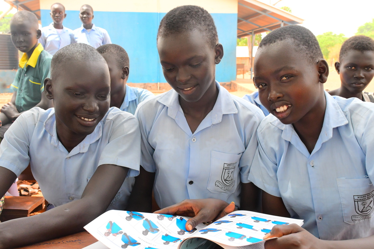 Auf dem Bild sind drei Mädchen im Südsudan zu sehen. Sie lächeln in die Kamera. Vor ihnen liegt ein Heft zur Gebärdensprache.