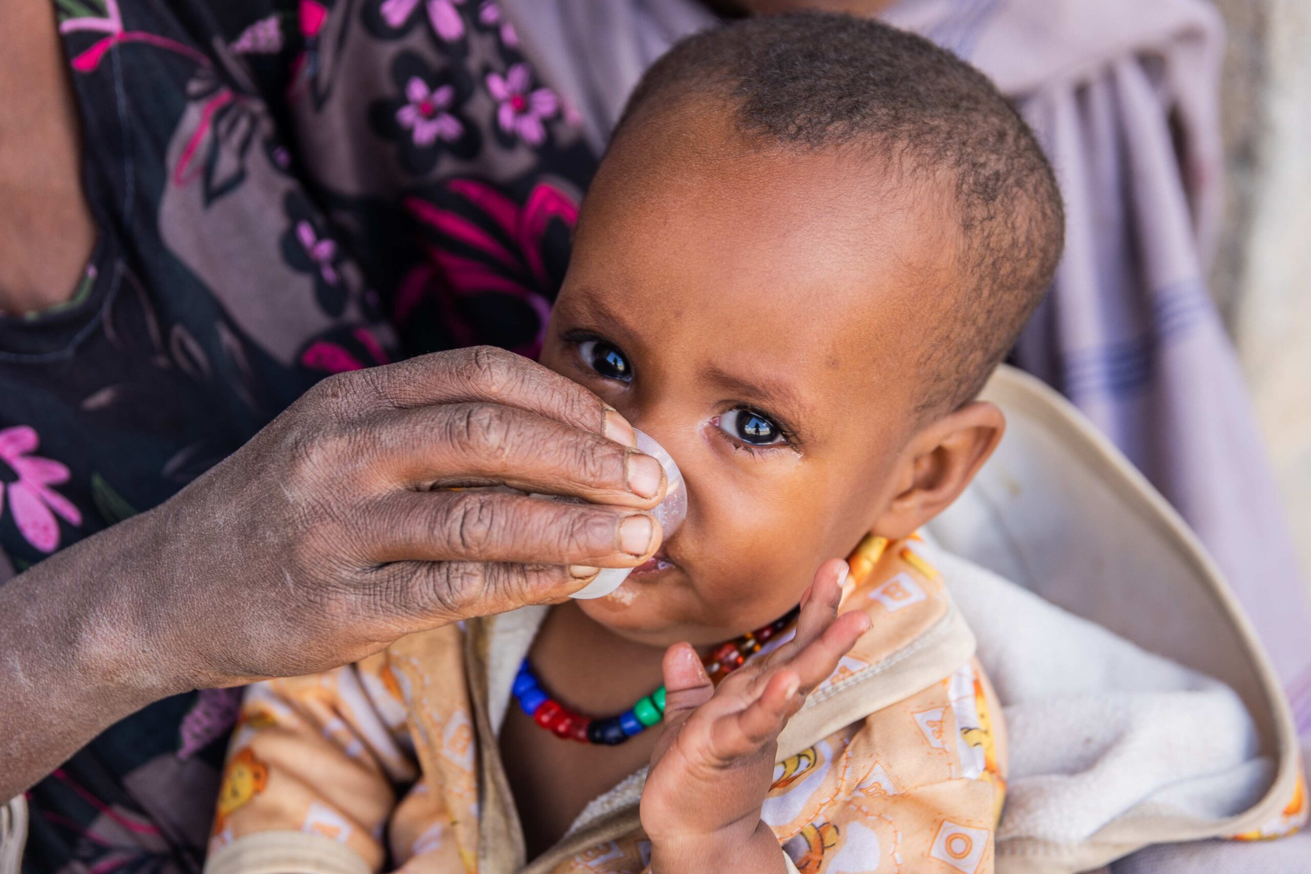 Ein Baby schaut in die Kamera, während es aus einem Plastikbecher Medizin trinkt.