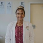 Eine schwarze Frau in einem weißen Kittel und einer roten Bluse blickt in die Kamera. Sie ist Kinderaugenärztin in einem Spital in Uganda.