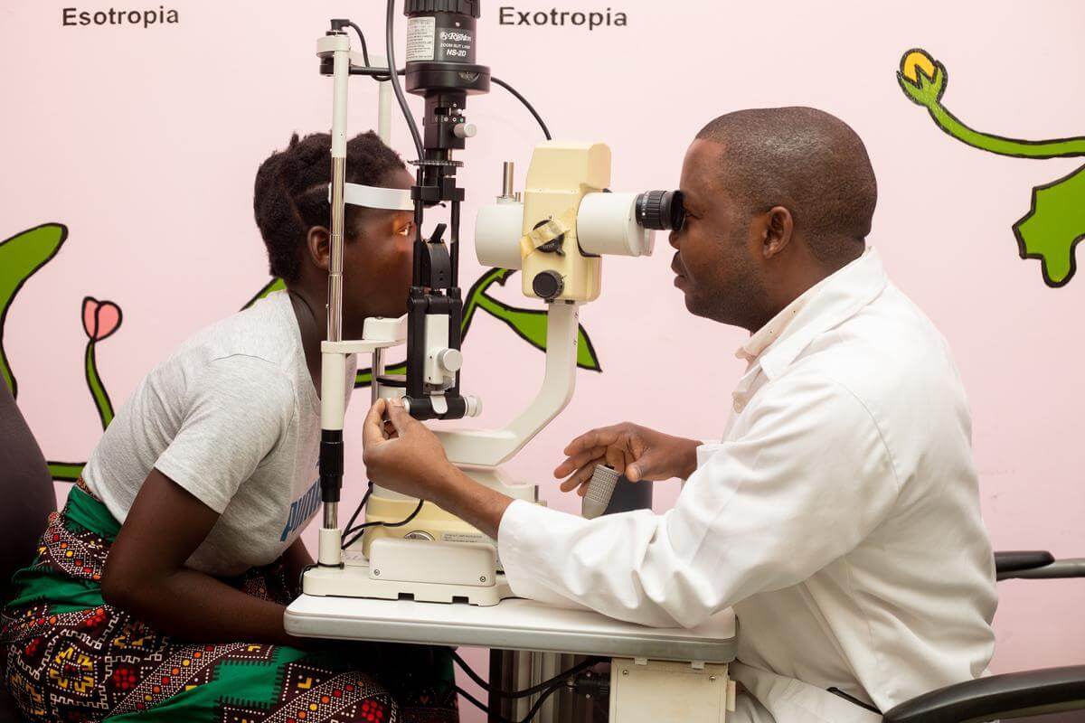 Auf dem Foto sind ein Augenarzt und seine junge Patientin zu sehen. Der Arzt trägt einen weißen Kittel, das Mädchen ein graues T-Shirt. Beide haben eine dunkle Hautfarbe.