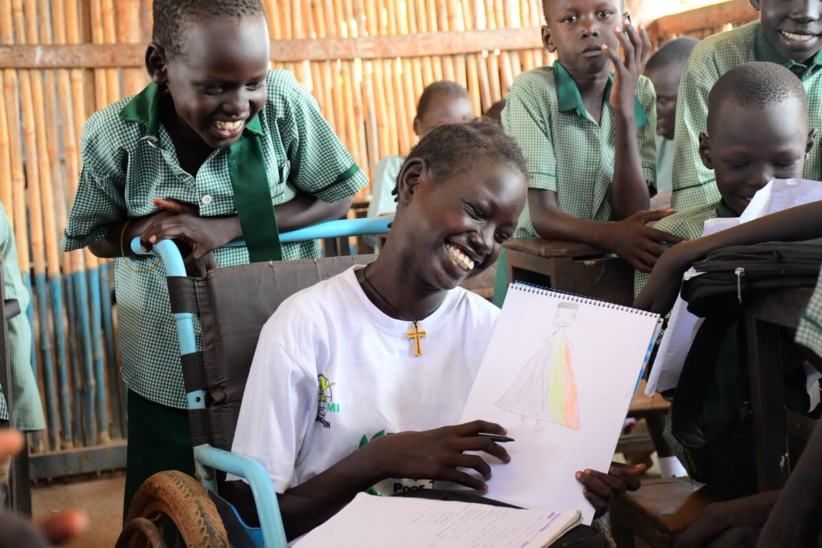 Ein Junge mit schwarzer Hautfarbe sitzt im Rollstuhl und zeigt eine Zeichnung in die Kamera. Er hat ein breites Grinsen im Gesicht. Um ihn herum stehen seine Klassenkollegen.