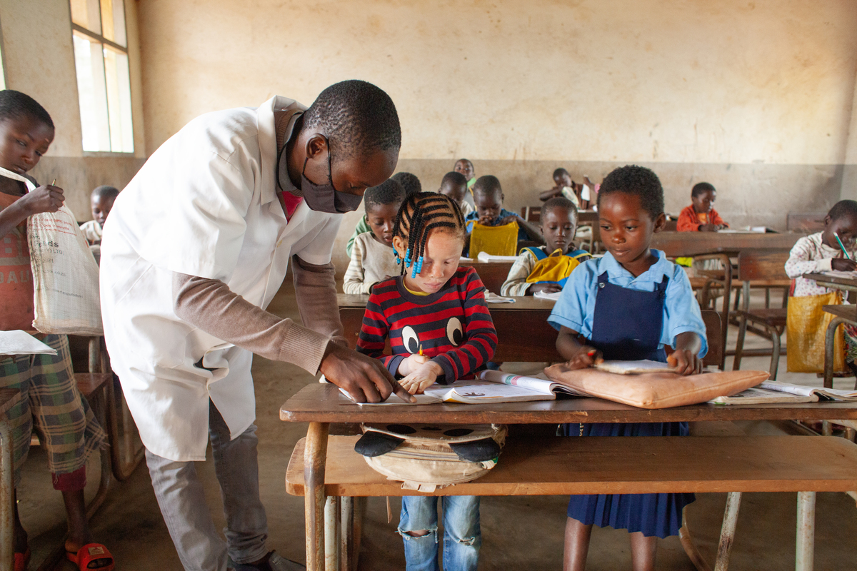 Die Szene auf dem Foto spielt in einem Gebäude in Mosambik. Es ist ein Lehrer mit schwarzer Hautfarbe zu sehen, der einem Mädchen mit Albinismus etwas in einem Schulheft zeigt.