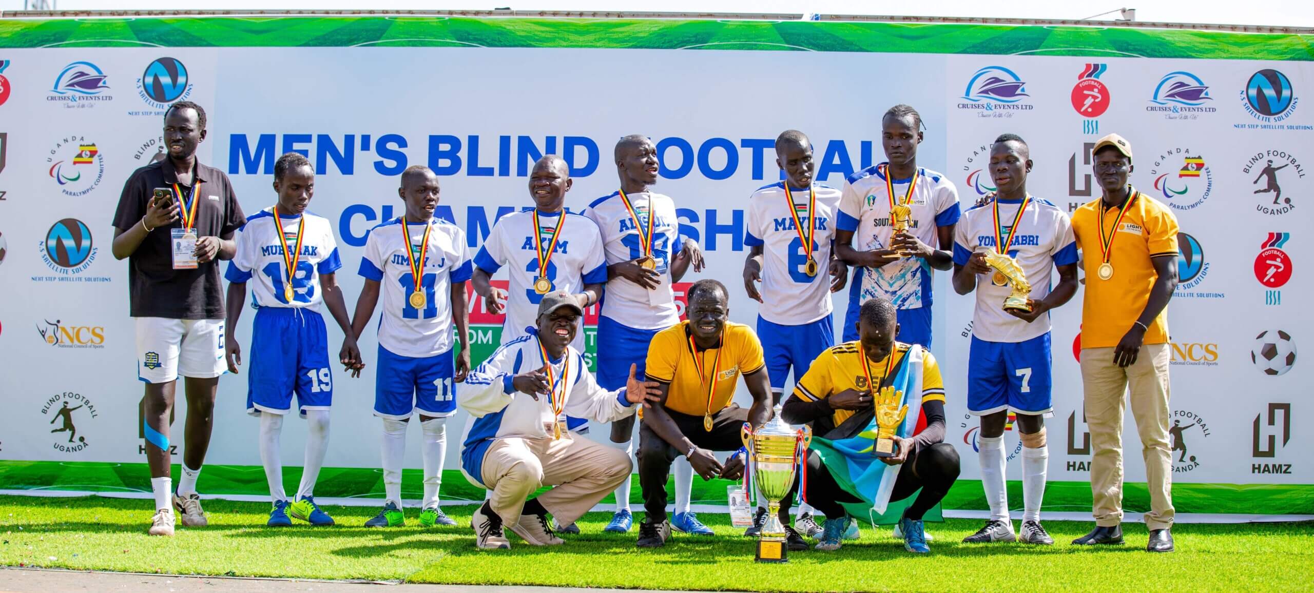 Eine Fußballmannschaft mit weiß-blauen Trikots posiert vor einer Foto-Wand. Sie tragen Medaillen und vorne steht ein goldener Pokal. Sie haben ein wichtiges Spiel gegen Uganda gewonnen.