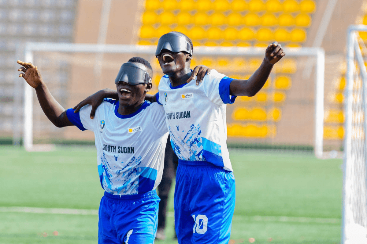 Zwei Blindenfußballspieler liegen sich den Armen und jubeln. Sie haben gerade das Spiel gegen Uganda gewonnen. Sie tragen ein weiß-blaues Trikot und schwarze Brillen. Im Hintergrund ist ein Fußballtor und eine Tribüne mit gelben Stühlen zu sehen.