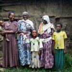 A family of six stands together. The three adult women wear long dresses and look at each other happily. One of them wears a blindfold from her past operation.