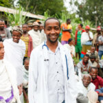 A doctor from Ethiopia in a white coat stands in a crowd and smiles at the camera.