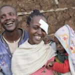 Husband, wife and child smiling and being happy over the fact that Tesfanesh (the wife) got an eye surgery and can see again. Tesfanesh is looking at the camera, smiling with an eye patch on her left eye. She is holding is standing in front of her husband, holding her child.