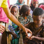 A boy from Ethiopia holds out both hands to get pink pills against the infectious eye disease trachoma. Behind him are other children waiting in line.