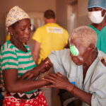 An elderly woman with a blindfold is trying to stand up. She is wearing a grey dress with press studs at the shoulders. A younger woman assists her to stand up.