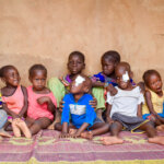 Many children with and without disabilities are sitting in front of a brown wall. They are all colourfully dressed and laughing.